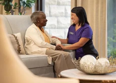 A smiling Amica caregiver sitting beside an elderly woman on a lounge sofa, holding her hands warmly in a bright, comfortable common area
