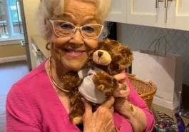 A cheerful elderly woman wearing glasses and a pink top smiles while holding a small teddy bear close to her