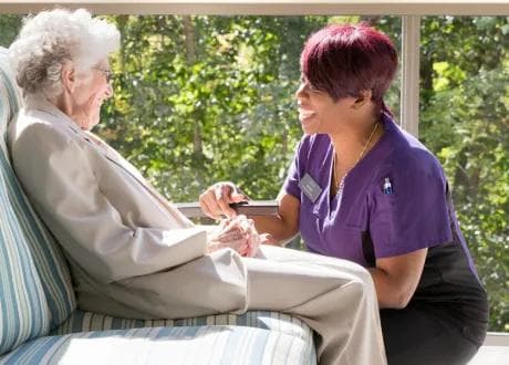 An Amica caregiver sits attentively beside an elderly woman near a window, holding her hands in a warm, engaged conversation