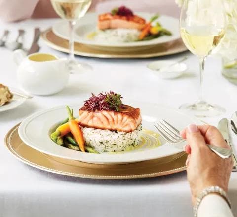 An elegantly plated salmon fillet serving on a bed of rice with asparagus and roasted carrots, garnished with greens, on a plate with wine glasses and cutlery on a white linen table