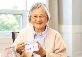 A smiling elderly woman wearing glasses holds up a small decorated card or artwork while seated near a bright window