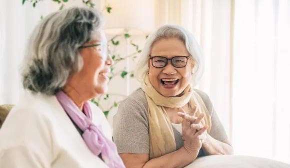 Two senior women sitting together near a bright window, laughing and enjoying a lively conversation