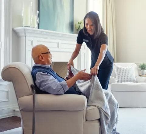 A smiling Amica caregiver draping a blanket over an elderly man seated comfortably in an armchair in a bright, cozy living room
