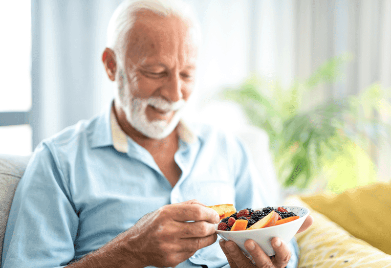 Man sits in a chair smiling, as he eats berries and a sliced peach from a bowl.