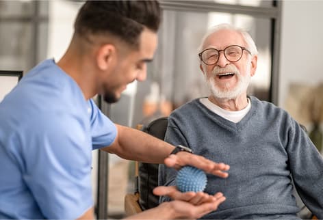 Mans sits and smiles with senior - Banner desktop Man shows senior how to use therapy ball between his hands.
