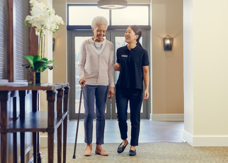 Lifestyle Shoot - Resident and Caregiver Walking - Cropped Female resident walking down hallway with support of cane and female caregiver